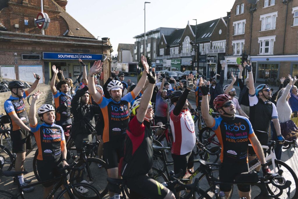 Cyclists taking part in Ride 1000 and their friends and family cheer and wave outside Southfields Undergound Station before setting off