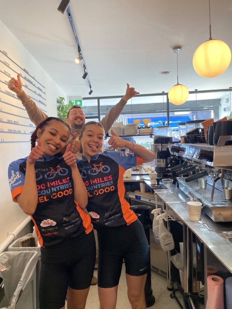 2 young people joining Ride 1000 and a Regenerate staff member posed with their thumbs up behind the counter in the Feel Good coffee shop, Southfields